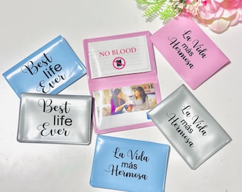 Puede incluir: Varios tarjeteros en tonos azul, plateado y rosa. Los tarjeteros tienen las frases "Best life Ever" y "La Vida más Hermosa" impresas en escritura negra. Un tarjetero está abierto, mostrando una foto en su interior.