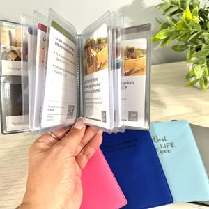 May include: A clear plastic booklet with several pages of printed text, held open by a hand. The booklet is in a clear plastic holder. Several colorful, small notebooks are visible below the booklet.