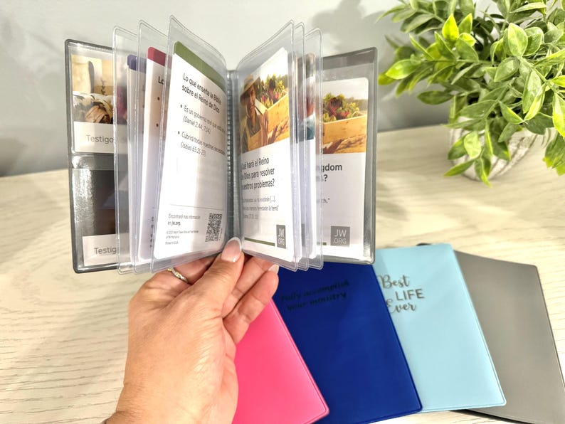May include: A clear plastic booklet with several pages of printed text, held open by a hand. The booklet is in a clear plastic holder. Several colorful, small notebooks are visible below the booklet.