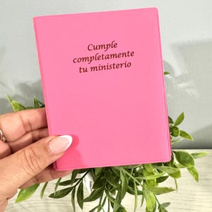 May include: A bright pink notebook with the text "Cumple completamente tu ministerio" in brown lettering. The notebook is held over green leafy sprigs.