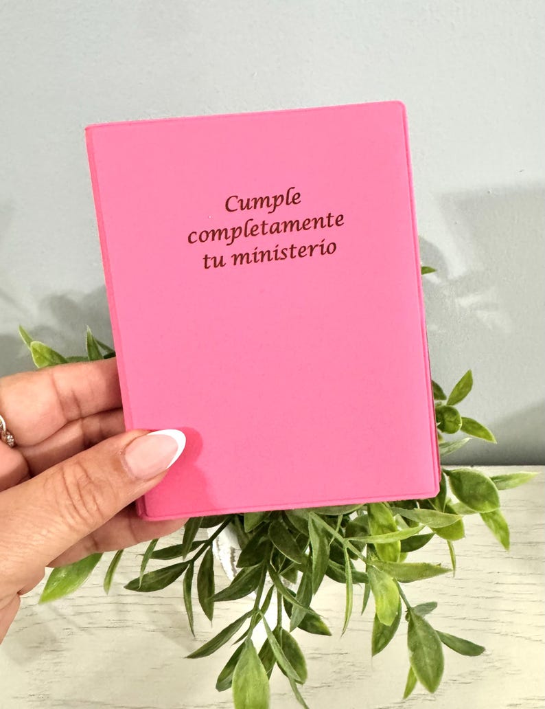 May include: A bright pink notebook with the text "Cumple completamente tu ministerio" in brown lettering. The notebook is held over green leafy sprigs.