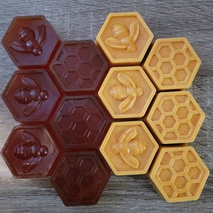 May include: A set of 12 hexagonal honey-themed soap bars. The soaps are in two colors: brown and yellow. The brown soaps have a honeycomb pattern and a bee design. The yellow soaps have a honeycomb pattern and a bee design.
