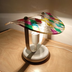 May include: A white table lamp with a colorful geometric glass shade. The shade is made of clear glass with a pattern of squares in pink, green, yellow, and blue. The lamp has a white base and a silver metal ring.