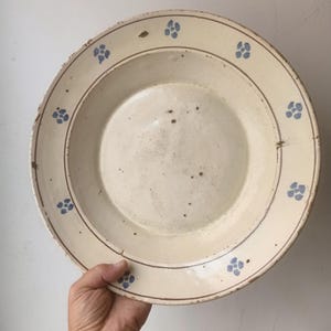 Puede incluir: Un plato redondo de cerámica con acabado esmaltado en color blanco roto, con un borde ancho decorado con un patrón repetitivo de pequeñas flores azules de cuatro pétalos y finas líneas circulares marrones, con signos de desgaste.