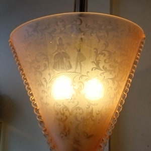 Puede incluir: Una lámpara colgante de vidrio rosa con un diseño grabado decorativo de dos figuras y motivos florales. La lámpara tiene un borde de cuentas y dos bombillas.