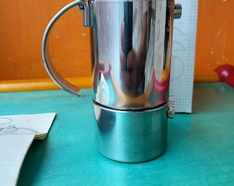 May include: A stainless steel stovetop espresso maker with a curved handle. The coffee pot has a cylindrical body and a screw-on base. A box with the text "1 tazza" is visible in the background.