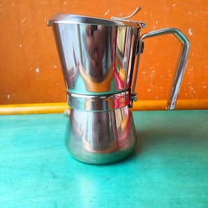 Puede incluir: Una cafetera espresso de acero inoxidable pulido con una cámara superior ancha y cónica, una base cilíndrica y un mango angular resistente. La tapa incluye una pequeña palanca metálica para abrirla y la superficie exterior es muy reflectante.