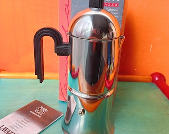 May include: A silver and black stovetop espresso maker with a black handle. The maker is sitting on a blue surface with a red and orange surface in the background. The box for the espresso maker is visible in the background. The box is white with black text that reads "CAMILLA AZZURRA Caffettiera  Espresso". A small instruction booklet is visible in the foreground. The booklet is white with black text that reads "LAVAZZA PRODUZIONE".