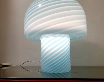 May include: A blue and white striped glass table lamp with a mushroom-shaped shade. The lamp is turned on and the light shines through the glass.