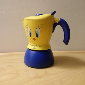Puede incluir: Cafetera Moka Express amarilla y azul con un diseño de Piolín. La cafetera tiene un mango azul y una base azul.