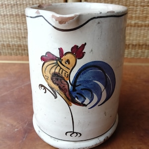 Peut inclure: Pichet en céramique blanche avec un motif de coq peint à la main. Le coq a un corps jaune, une queue bleue et une crête rouge. Le pichet a un aspect légèrement vieilli avec un bord et une base foncés.