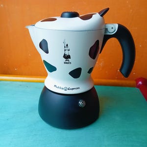 May include: A black and white espresso maker with a cow-like pattern. The coffee maker has a black handle and a small logo of a man with a mustache. The text "Mukka Express" is printed on the base.
