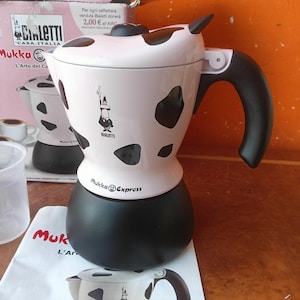 Könnte beinhalten: Ein Bialetti Mukka Express Herdkocher mit einem weißen Oberteil mit schwarzen Kuhflecken, einem schwarzen Unterteil und einem schwarzen Griff. Das Markenlogo und der Schriftzug Mukka Express sind auf der Vorderseite des weißen Teils aufgedruckt.