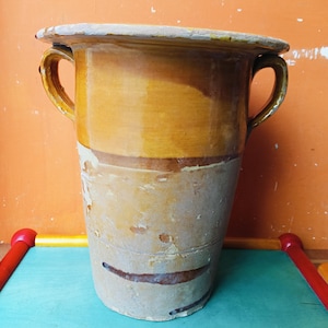 Könnte beinhalten: Eine hohe, antike Keramikvase mit breitem Rand und zwei gebogenen Griffen. Die Vase hat ein zweifarbiges Farbschema, mit einer hellbraunen unteren Hälfte und einer dunkleren, goldgelben oberen Hälfte. Die Vase hat ein rustikales, gealtertes Aussehen.