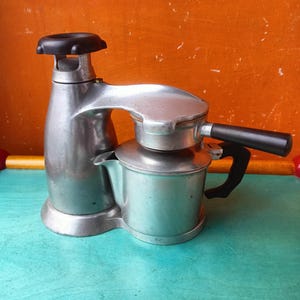 Puede incluir: Una cafetera espresso vintage de aluminio para estufa que presenta una caldera cilíndrica alta con una perilla negra superior, una cámara de preparación conectada con un mango lateral negro y un mango negro a juego en el cuerpo principal.