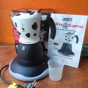 Pode incluir: Uma cafeteira elétrica Bialetti Mukka Express com a parte superior com estampa de vaca em branco e preto, base preta e alça preta. Inclui um cabo de alimentação, um copo medidor translúcido, um manual de instruções e a caixa original.
