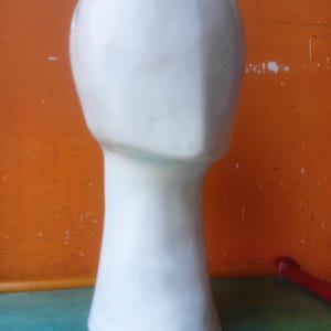 Könnte beinhalten: Ein weißer Keramik-Mannequinkopf mit glatter, matter Oberfläche. Der Kopf ist oval geformt mit langem Hals und flachem Sockel. Der Hintergrund ist eine leuchtend orange Wand.