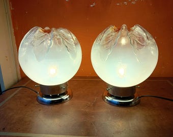 May include: Two matching table lamps. Each lamp features a frosted glass globe with a clear, leaf-like design at the top. The lamps sit on a chrome base and are illuminated, casting a soft glow. The lamps are connected to a power source.