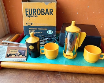 Pode incluir: Um conjunto de cafeteira Eurobar Express vintage. O conjunto inclui uma cafeteira amarela e prateada, duas chávenas amarelas, um recipiente preto e amarelo e uma caixa com o logótipo Eurobar. Desenhado para viagens e preparação rápida de café.