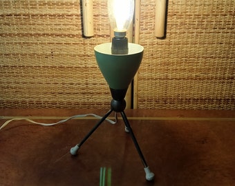 May include: A mid-century modern table lamp with a green ceramic shade and a black metal tripod base. The lamp has a clear light bulb with a warm white glow.