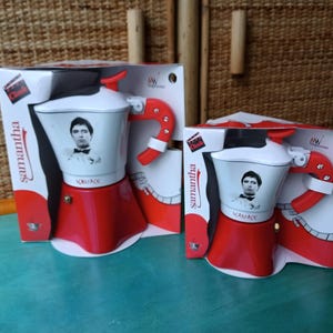Puede incluir: Dos cafeteras de cerámica roja y blanca con una imagen en blanco y negro de un hombre en la parte delantera. Las cafeteras están en su embalaje original, que es rojo y blanco con las palabras "Samantha" y "Clack" impresas en él.
