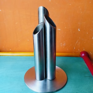 May include: A modern, brushed metal sculpture featuring three cylindrical tubes of varying heights, joined at the base. The sculpture has a circular base and a polished, reflective surface. The design is minimalist and contemporary.