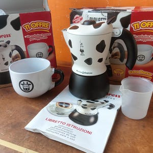 Op de afbeelding: Een Bialetti Mukka Express fornuis-cappuccinomaker met een wit en zwart koeienvlekkenpatroon, een zwarte basis en een zwarte handgreep. Inclusief een witte keramische mok met een koeiengezichtlogo, een doorzichtige plastic maatbeker en een handleiding.