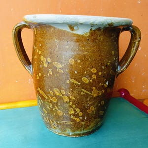 Könnte beinhalten: Eine braune Keramikvase mit zwei Henkeln und einer weiten Öffnung. Die Vase hat ein gesprenkeltes Design mit gelben und braunen Flecken. Der Rand ist weiß. Die Vase ist ein rustikales, handgefertigtes Stück.