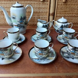 Puede incluir: Un juego de té japonés vintage con fondo blanco y diseños florales y paisajísticos azules y dorados. El juego incluye una tetera, una jarra de crema, un azucarero y seis tazas de té y platillos.