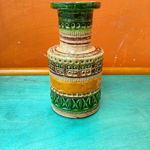 May include: A small, green, yellow and brown ceramic vase with a geometric pattern. The vase has a narrow neck and a wide base.