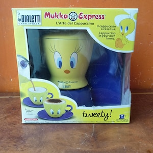 Könnte beinhalten: Ein gelb-blauer Bialetti Mukka Express Cappuccino-Bereiter in der Originalverpackung. Die Schachtel zeigt Tweety-Grafiken, den Schriftzug Mukka Express und Bilder von zwei gelben Tassen auf violetten Untertassen. Kapazität für 2 Tassen.