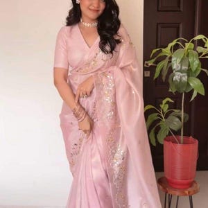 Puede incluir: Un sari de color rosa claro que presenta una blusa con cuello en V de manga corta y una falda a juego con una caída ligera y transparente. La tela está adornada con intrincados bordados florales en oro y plata a lo largo de los bordes.