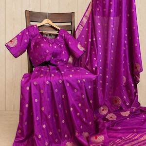 May include: A vibrant purple saree featuring a matching short-sleeved blouse. The fabric is adorned with small gold polka dots and larger circular motifs with red centers. The border displays horizontal gold stripes and intricate floral patterns.