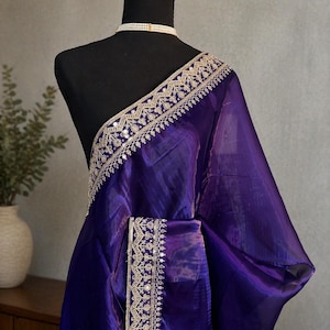 Puede incluir: Un sari transparente de color morado intenso con un borde ancho y ornamentado que presenta intrincados bordados dorados y pequeños detalles de espejos reflectantes. La tela tiene un brillo sutil y está sobre un maniquí negro con un collar de perlas.