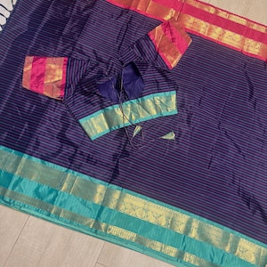 May include: A dark blue saree featuring thin horizontal stripes and a wide teal border with gold zari patterns. The matching blouse has short sleeves with pink and gold bands, a deep neckline, and thin tie-up strings at the center back.