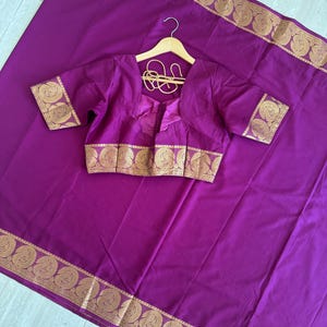 May include: A vibrant purple saree paired with a matching short-sleeved blouse, both featuring wide gold borders with intricate paisley patterns. The blouse includes a square neckline and thin gold tie-up strings at the back.