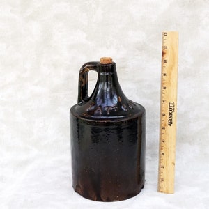 May include: A brown stoneware jug with a cork stopper and a handle. The jug is approximately 9 inches tall.