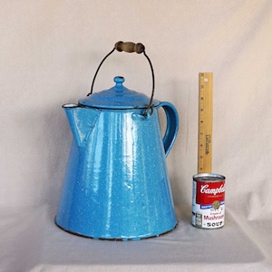 May include: A blue enamel camping coffee pot with a wooden handle and a speckled finish. The pot has a spout and a lid. The pot is sitting on a white surface next to a ruler and a can of soup.