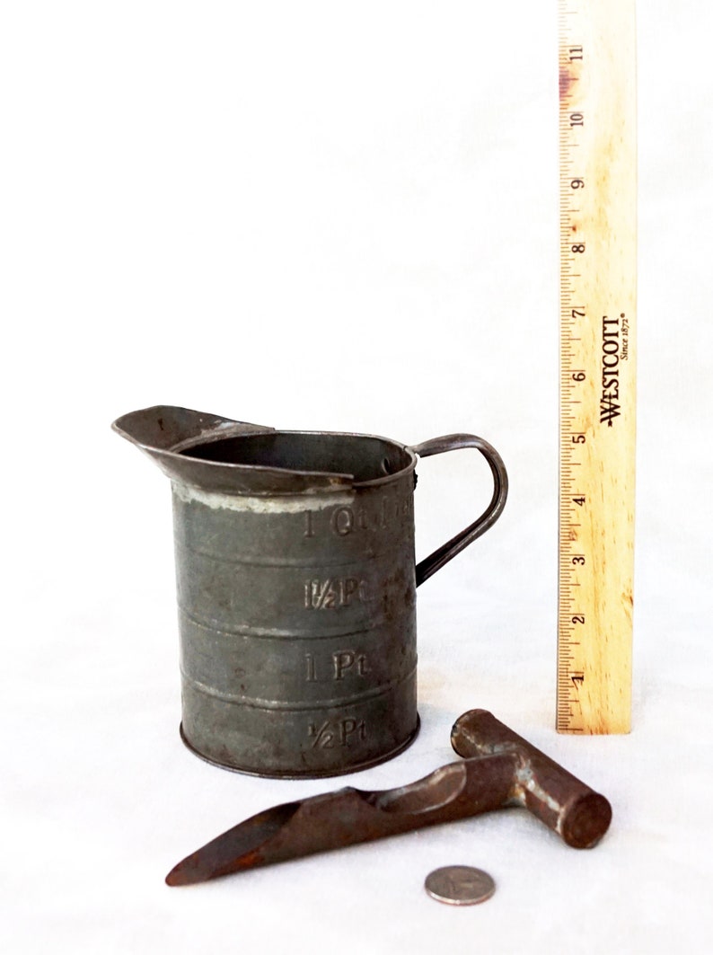 May include: A vintage metal measuring cup with a handle, a metal tool with a handle, and a silver coin. The measuring cup has markings for 1/2 pint, 1 pint, 1/2 quart, and 1 quart.