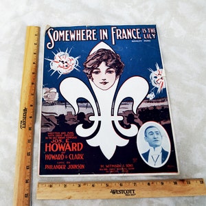 May include: A vintage sheet music cover for the song "Somewhere in France is the Lily". The cover features a white fleur-de-lis with a woman's face in the center. The background is blue with explosions and soldiers in the distance. The text "Written and sung with great success by the well known composer Jos. E. Howard of Howard & Clark Lyric by Philander Johnson" is printed on the cover.
