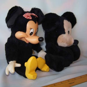May include: Two plush toys, Mickey Mouse and Minnie Mouse, are sitting side-by-side. Mickey is black with a tan face and large ears. Minnie is black with a red bow and a tan face. Both have yellow shoes.