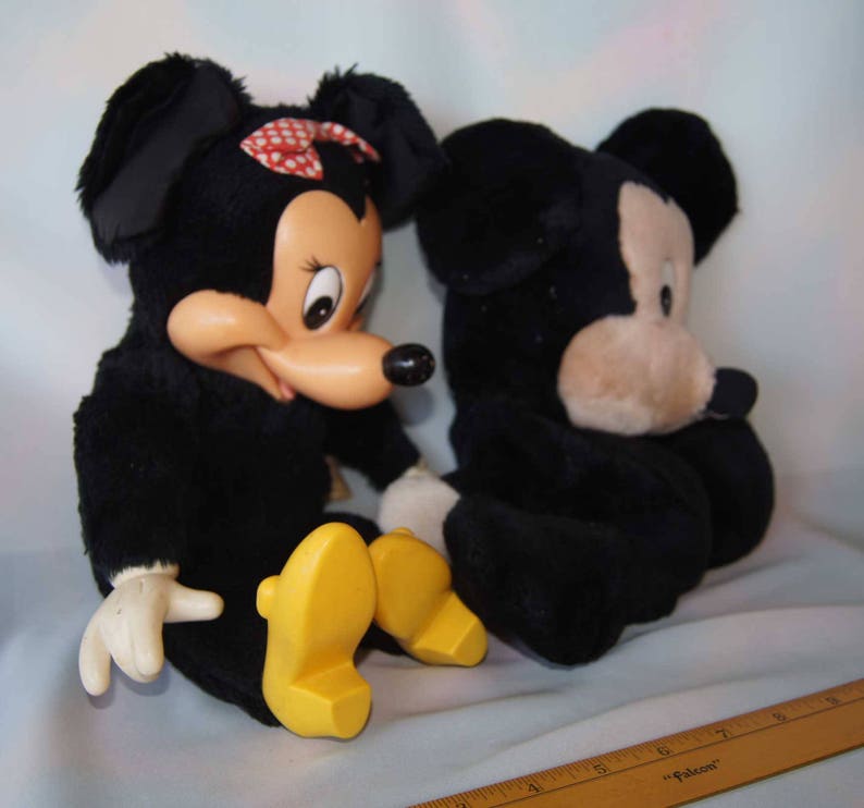 May include: Two plush toys, Mickey Mouse and Minnie Mouse, are sitting side-by-side. Mickey is black with a tan face and large ears. Minnie is black with a red bow and a tan face. Both have yellow shoes.