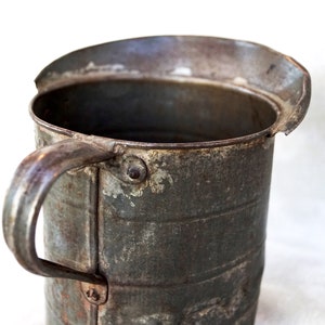 May include: A vintage metal measuring cup with a dented rim and a handle. The cup is made of galvanized metal and has a rustic, aged appearance.