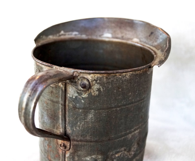 May include: A vintage metal measuring cup with a dented rim and a handle. The cup is made of galvanized metal and has a rustic, aged appearance.