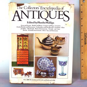 May include: The Collectors' Encyclopedia of Antiques book cover featuring a silver teapot, a silver creamer, a silver sugar bowl, a wooden cabinet with drawers, a blue and white porcelain plate, a blue and white porcelain vase, a blue and white glass goblet, a miniature horse-drawn carriage, and a miniature silver teapot and creamer. The book cover also includes the text "The Collectors' Encyclopedia of Antiques" and "Edited by Phoebe Phillips".