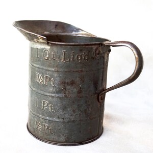 May include: A vintage metal measuring cup with a handle. The cup is marked with measurements in quarts and pints: 1 Qt. Liq'd, 1 1/2 Pt, 1 Pt, 1/2 Pt.