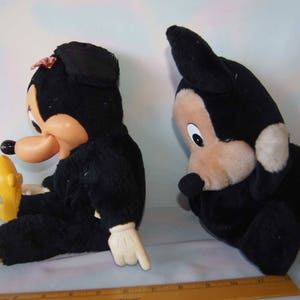 May include: Two plush toys, one black Mickey Mouse and one black Minnie Mouse, are sitting on a white surface. Mickey Mouse is wearing a red bow tie and Minnie Mouse is wearing a red bow.