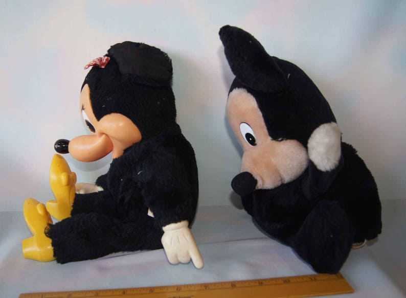 May include: Two plush toys, one black Mickey Mouse and one black Minnie Mouse, are sitting on a white surface. Mickey Mouse is wearing a red bow tie and Minnie Mouse is wearing a red bow.