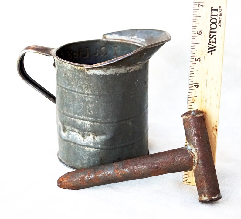 May include: A vintage metal measuring cup with a handle and a rusty metal tool with a T-shaped handle. The measuring cup is marked "1 Qt. Liquid".