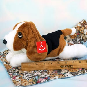 May include: A plush basset hound dog toy with brown, white, and black fur, lying on a patterned fabric. The toy has a red tag with a smiling face. A wooden ruler is next to the toy.